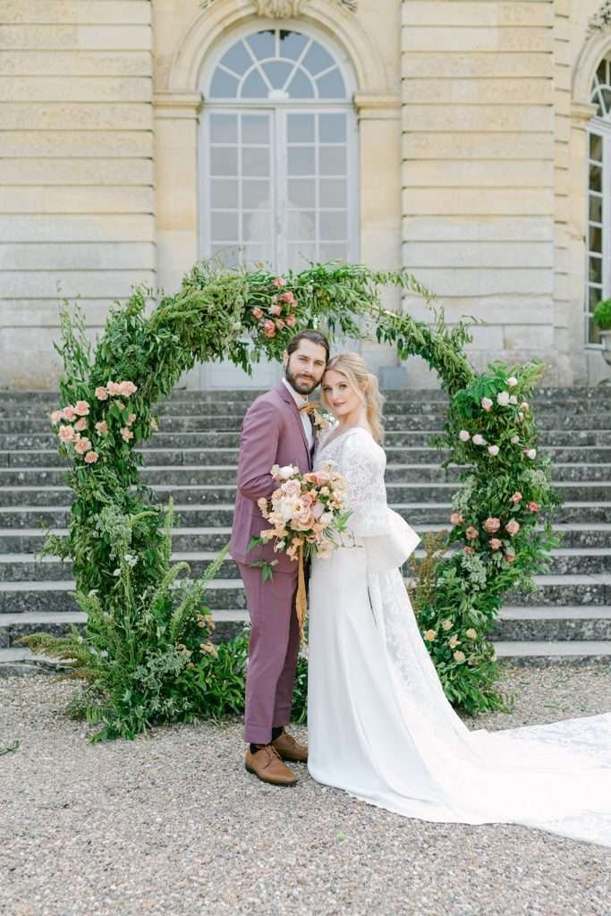 groom in a maroon suit and bride in a white lace gown standing with temples touching in front of a large greenery arch at the Chateau de Champlatreux Wedding venue