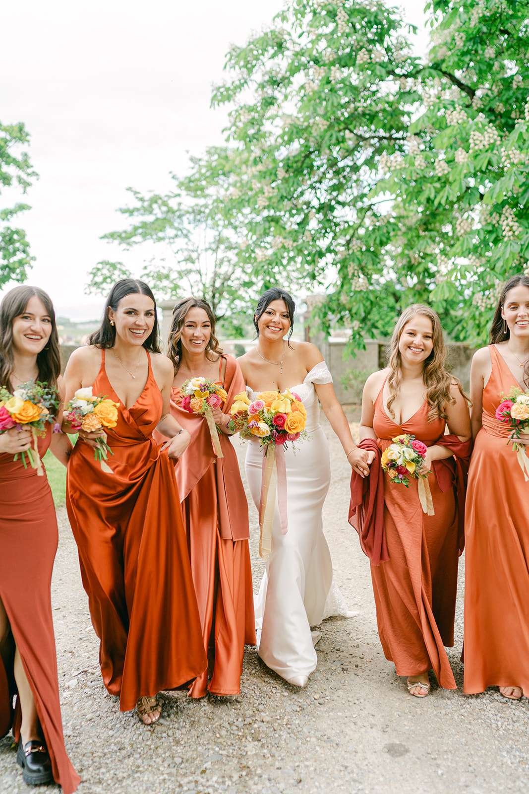 A bride smiles while walking the grounds of Château de Luins with her bridesmaids in orange dresses all holding bouquets