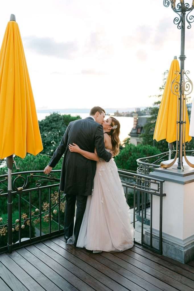 A bride and groom kiss on the balcony overlooking the lake at their Grand Hotel Suisse Majestic wedding