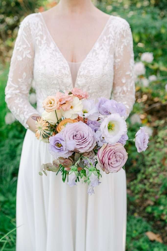 A bride stands in a garden holding her purple and pink bouquet in a lace dress