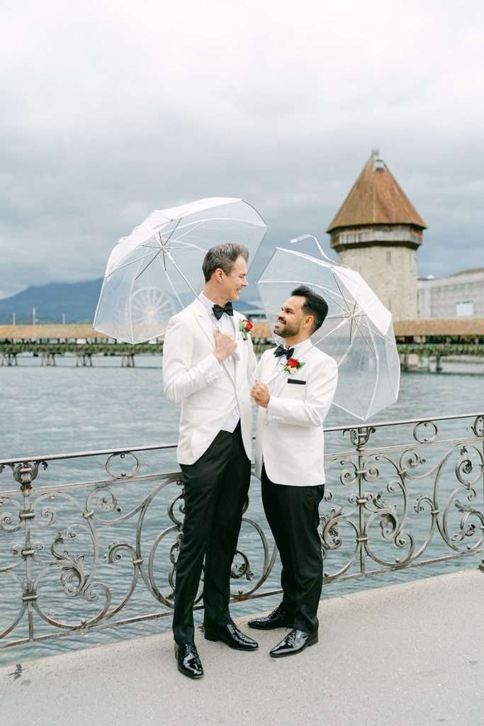 Two grooms in white jacket tuxes stand under clear umbrellas by the water against an ornate railing smiling at each other
