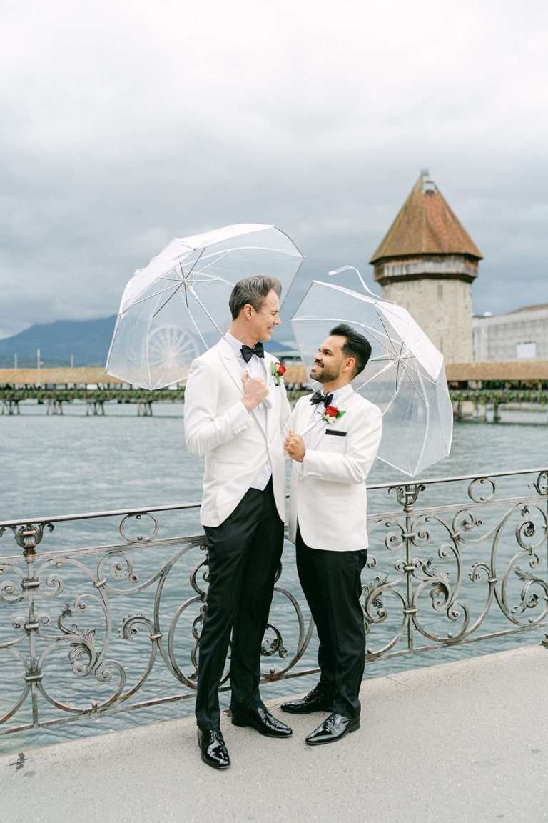 Two grooms in white jacket tuxes stand under clear umbrellas by the water against an ornate railing smiling at each other