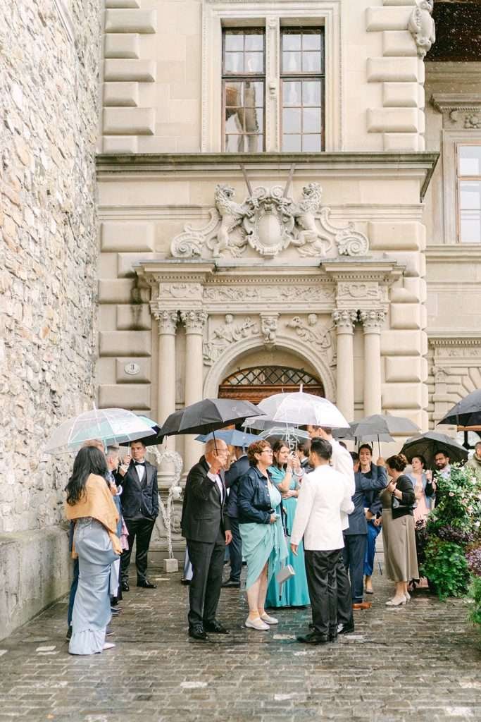 A group of wedding guests stand with umbrellas in the gardens of Beau Sejour Lucerne Switzerland