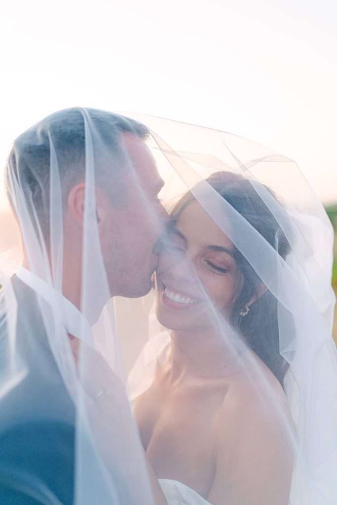 A groom kisses the bride on the cheek as she smiles when hiding under the veil at sunset during their Beau Sejour Lucerne Switzerland wedding
