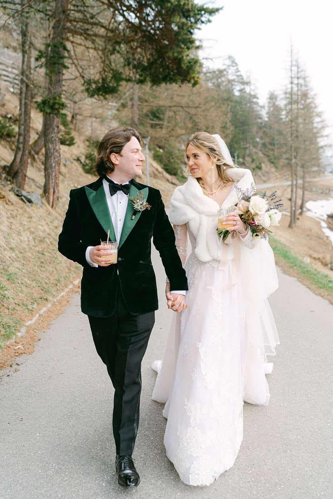 Newlyweds hold hands and smile at each other while holding drinks walking a mountain sidewalk