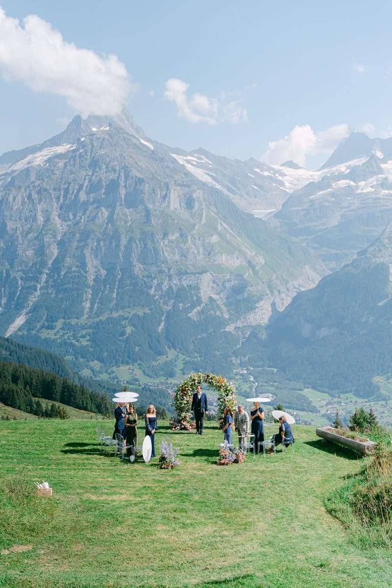 A look down at a micro wedding on the edge of a cliff with guests holding parasols and a floral arch